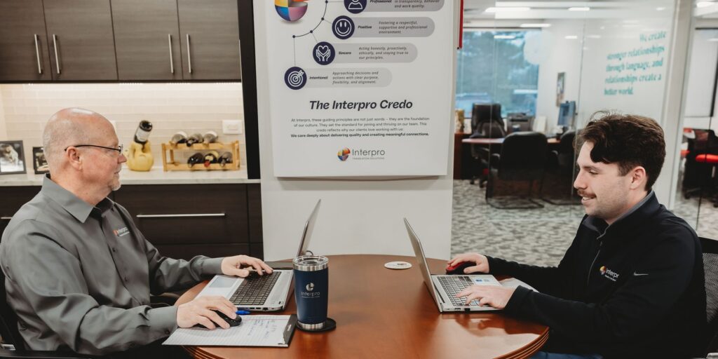 Two Interpro team members sitting at a conference table with laptops open, working in front of a company values display titled "The Interpro Credo"