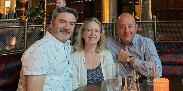 Sam Somerville, her husband, and Ralph Strozz sitting together at a restaurant table, smiling at the camera.